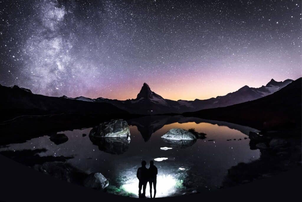 A couple stood in front of Stellisee lake at night time under the Milky Way. One reason why Zermatt is worth visiting, is for this stunning Astrophotography. One of the many things to do here.