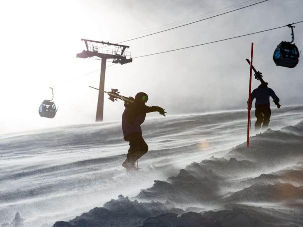 Skiers walking up a slope carrying their skis with gondolas overhead.