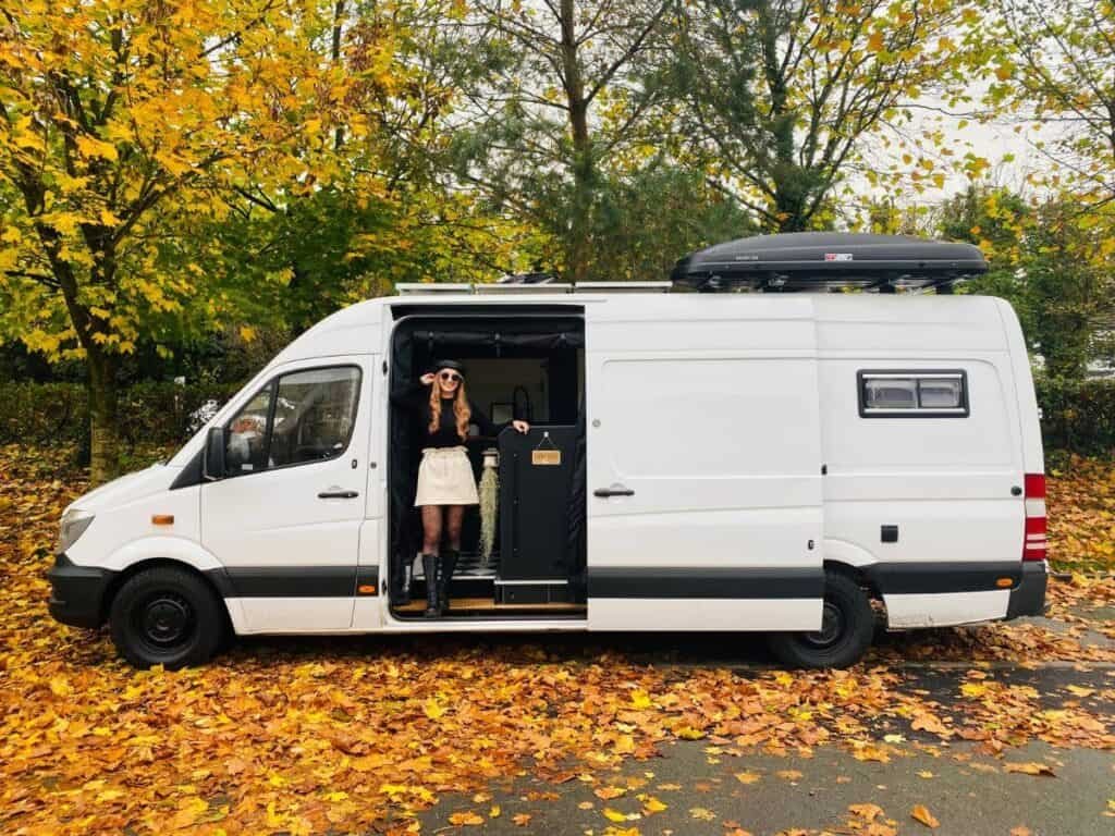 Vanlife parkup Scotland. A woman stood at the sliding door of her campervan during Autumn, surrounded by Autumn leaves.
