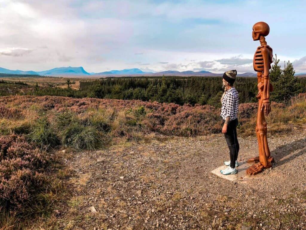 A skeleton sculpture in Borgie Forest named The Unknown, with a woman stood in front looking out at the same view over the surrounding landscape. A great location to add to your NC500 Itinerary.