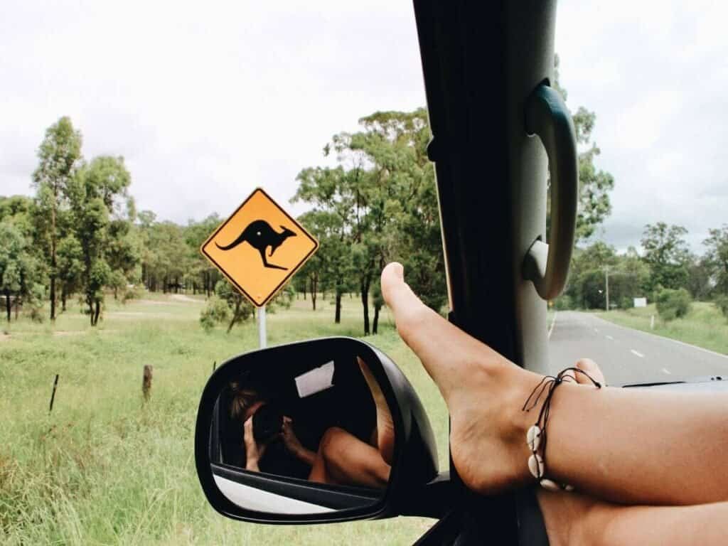 Woman with feet hanging out of the window of her vehicle, while passing a road sign for a kangaroo, in Australia.