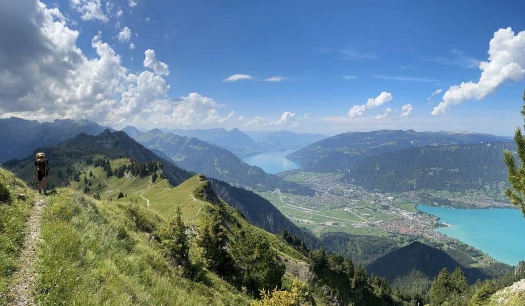 A hiker aproaching to the left of wide sweeping views of Interlaken, Lake Thun and Lake Brienz. This is vanlife in Switzerland.