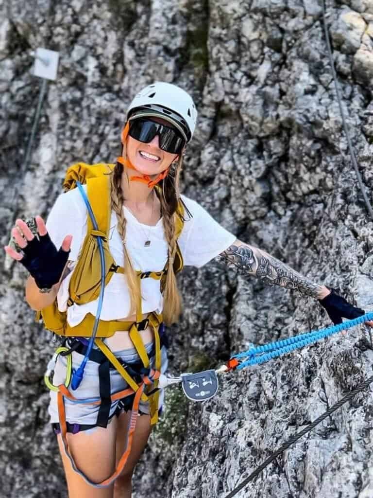 A woman smiling and waving in full via ferrata kit, attached to some rocks.