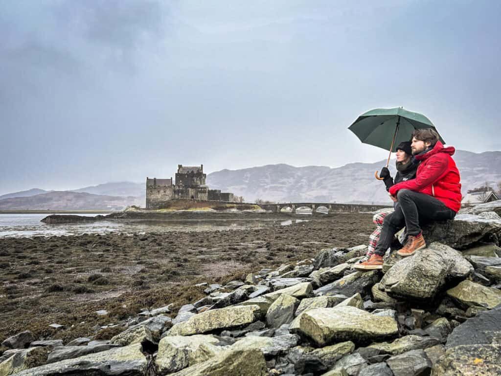 Man and woman sat on a rock on a moody weather day at Eileen Donan Castle, Scotland.